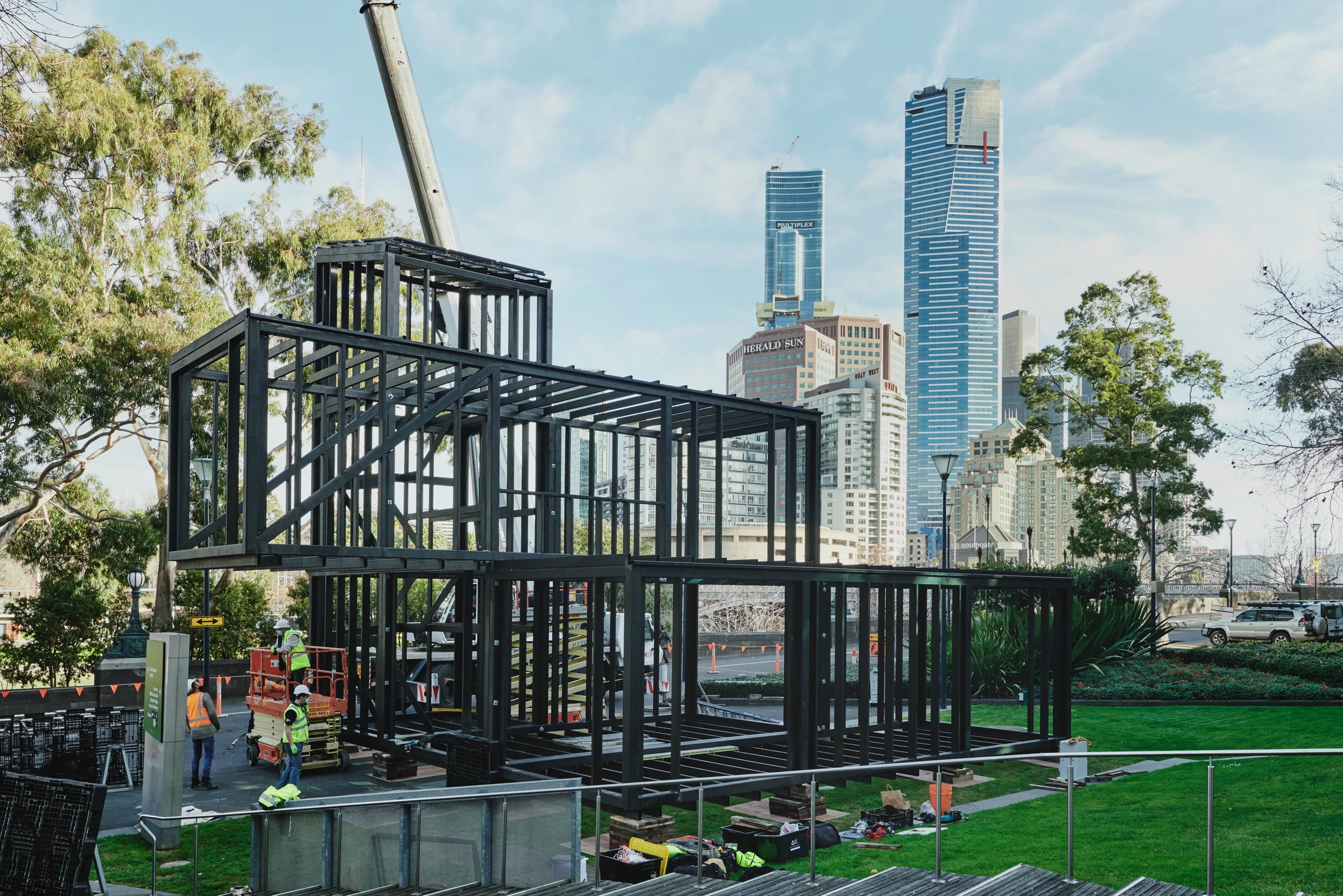 A black steel-frame modular building under construction in an urban park setting with city skyline visible