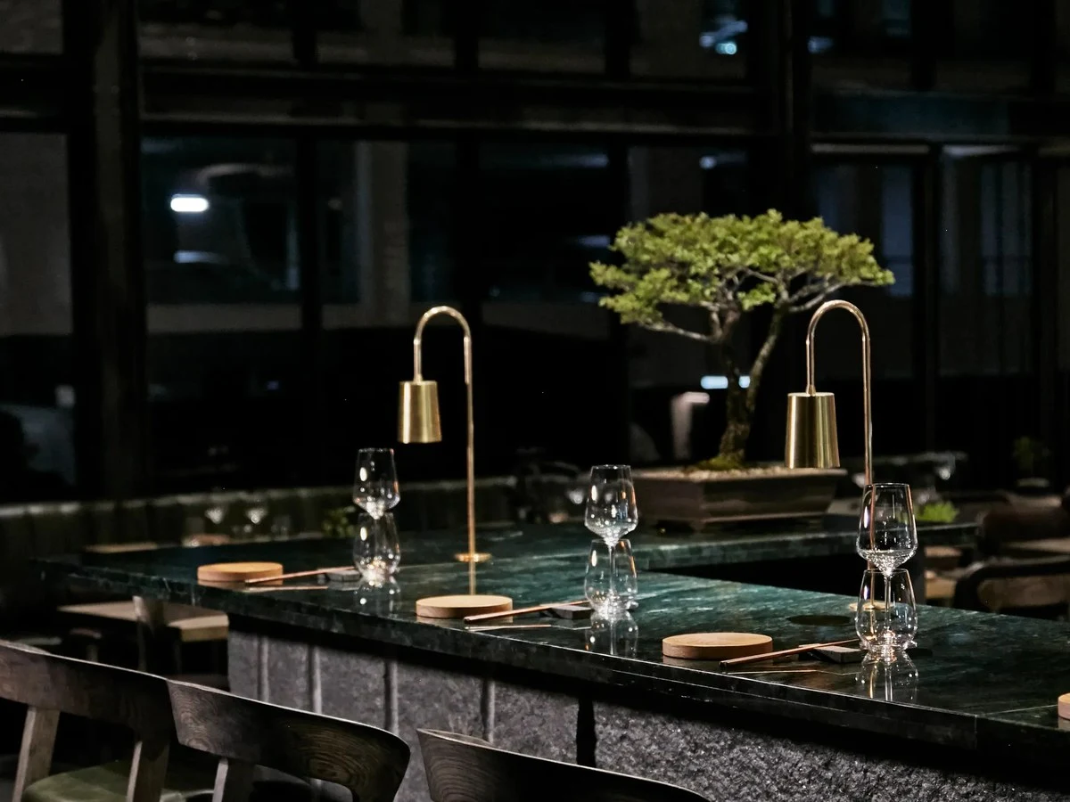 A dark, elegant bar interior at FYN Restaurant, Cape Town, featuring a deep green marble counter, brass arc lamps, crystal wine glasses, and a bonsai