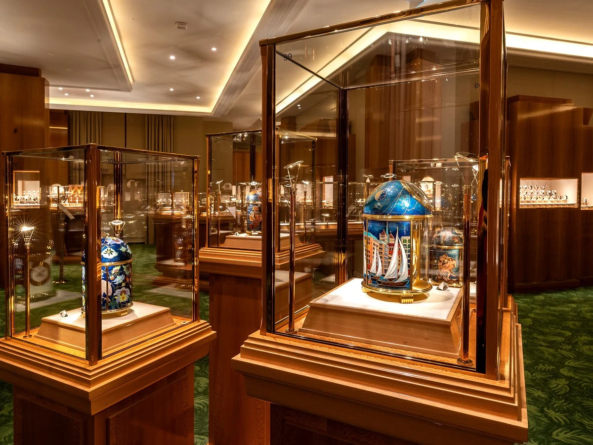 Interior view of a luxury museum or gallery space with multiple glass display cases containing ornamental objects, featuring warm lighting, wooden paneling, and a green patterned carpet.