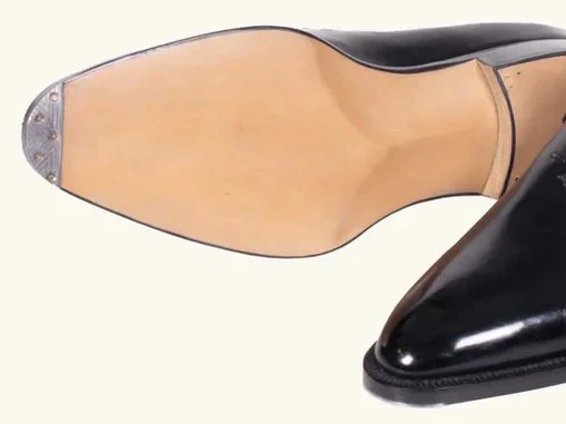 A pair of black leather Oxford dress shoes, one upright with a wooden shoe tree and the other inverted showing its leather sole.
