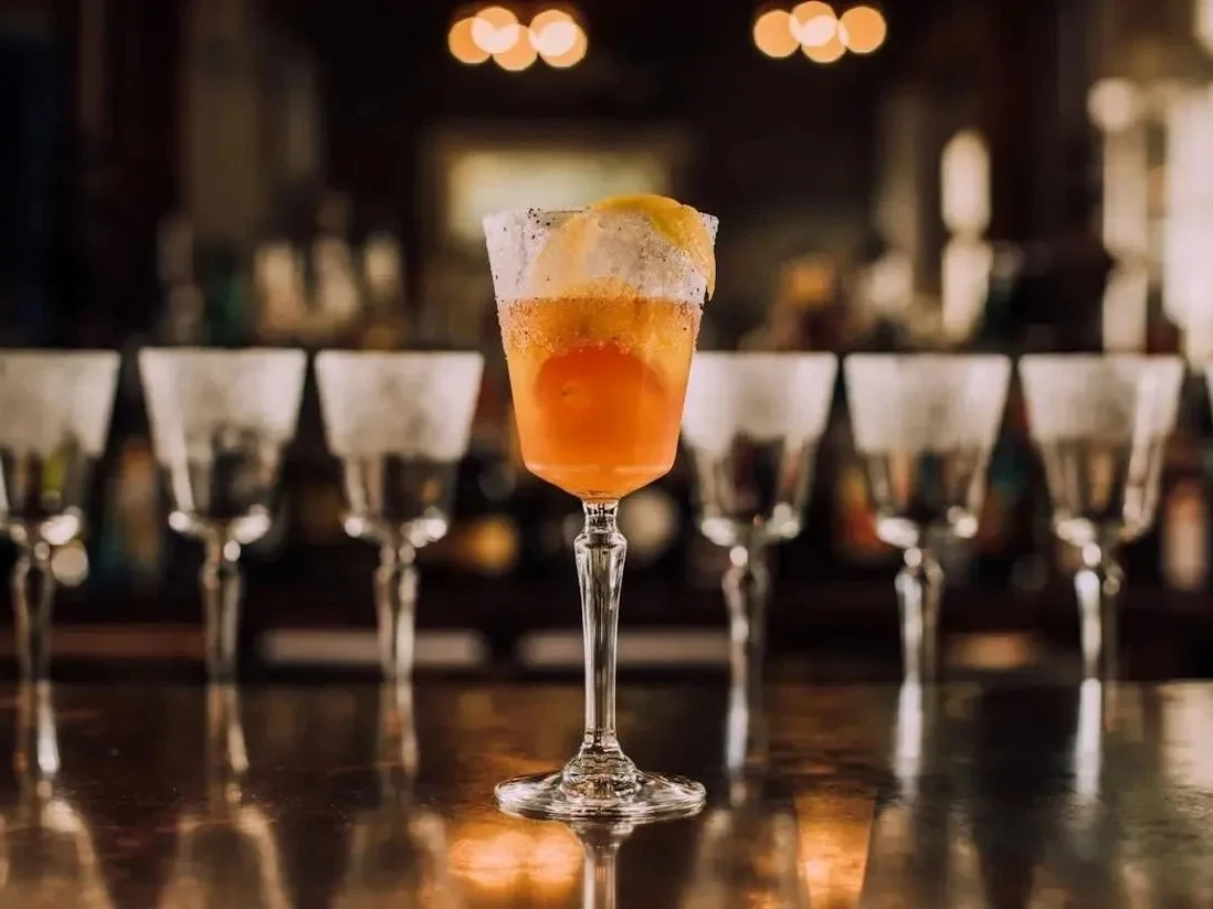A craft cocktail in an elegant stemmed glass with orange liquid, foam top, and citrus garnish, photographed in a bar setting with blurred bottles and 