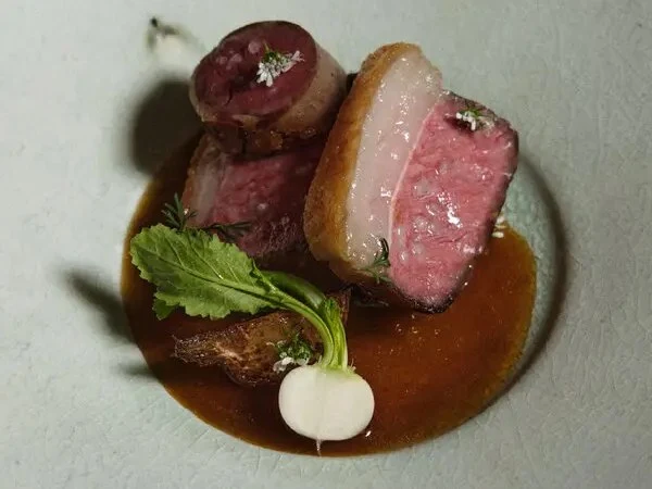 A white plate holds sliced pink meat, a small white turnip with green leaves, and dark brown sauce.