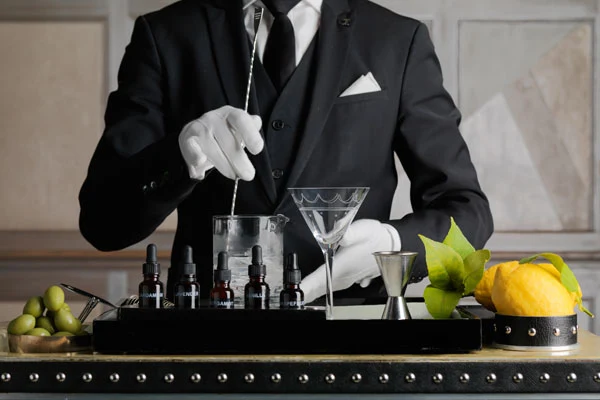A bartender wearing a black suit and white gloves stirs a cocktail with fresh lemons and limes visible on the bar counter.