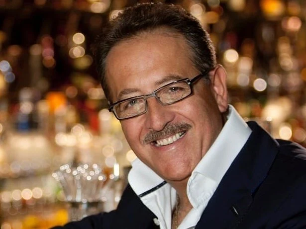 A smiling man in a dark suit and glasses leans on a marble bar, with a blurred backdrop of liquor bottles and warm lights.