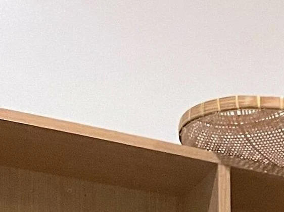 Close-up architectural detail showing the top edge of a wooden shelving unit or counter and a woven rattan/bamboo pendant lamp shade, with no person.