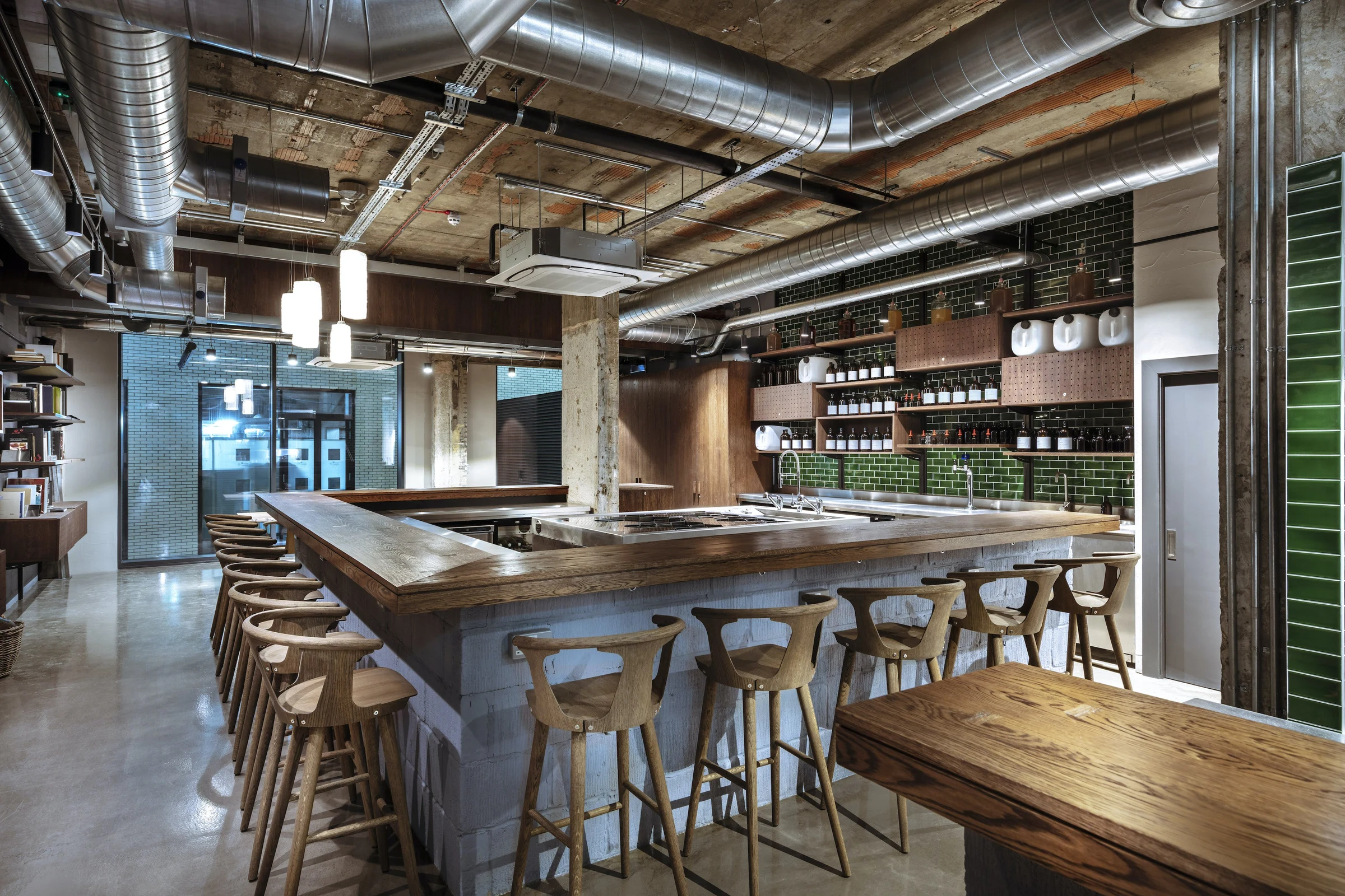 Interior of a contemporary bar with a large wooden counter, wooden bar stools, exposed ceiling with metal ducts, brick walls, and shelves of bottles behind the counter.
