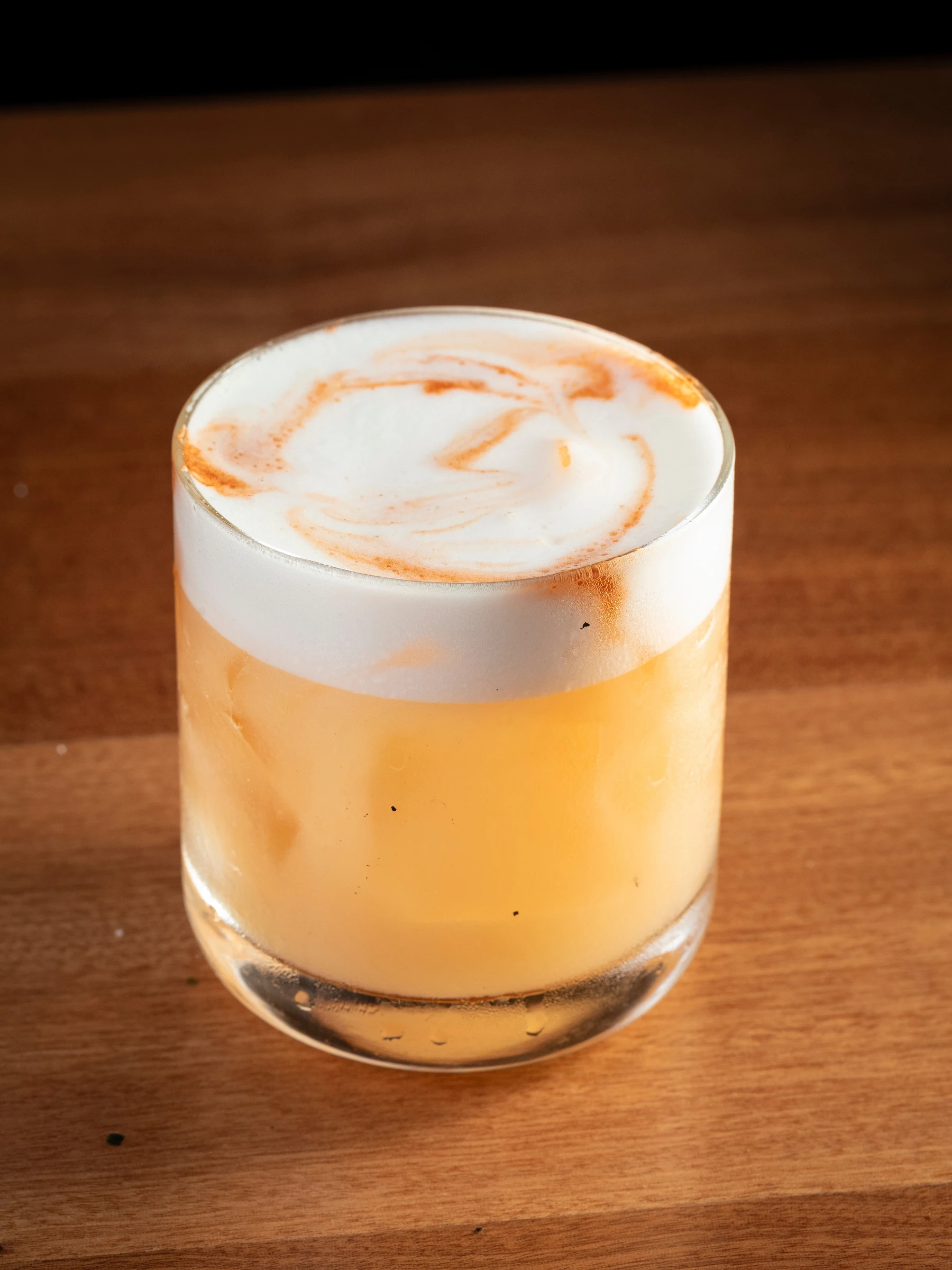 A cocktail in a rocks glass with a layered presentation, golden liquid base topped with white foam and decorative swirl pattern, identified by the sourc