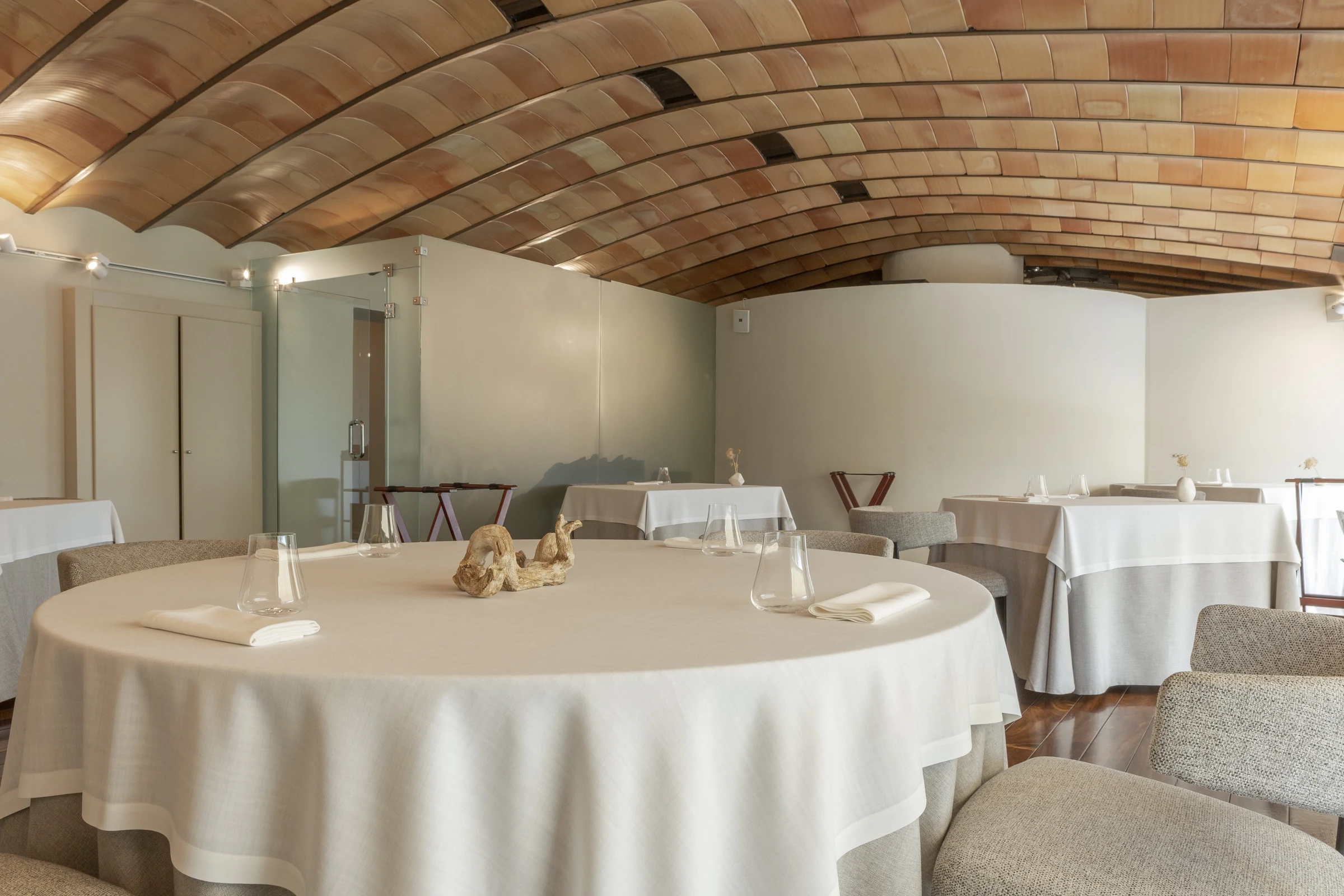 An upscale fine-dining restaurant interior with a distinctive curved wooden-tile ceiling, white-tablecloth tables, neutral upholstered seating, and mi