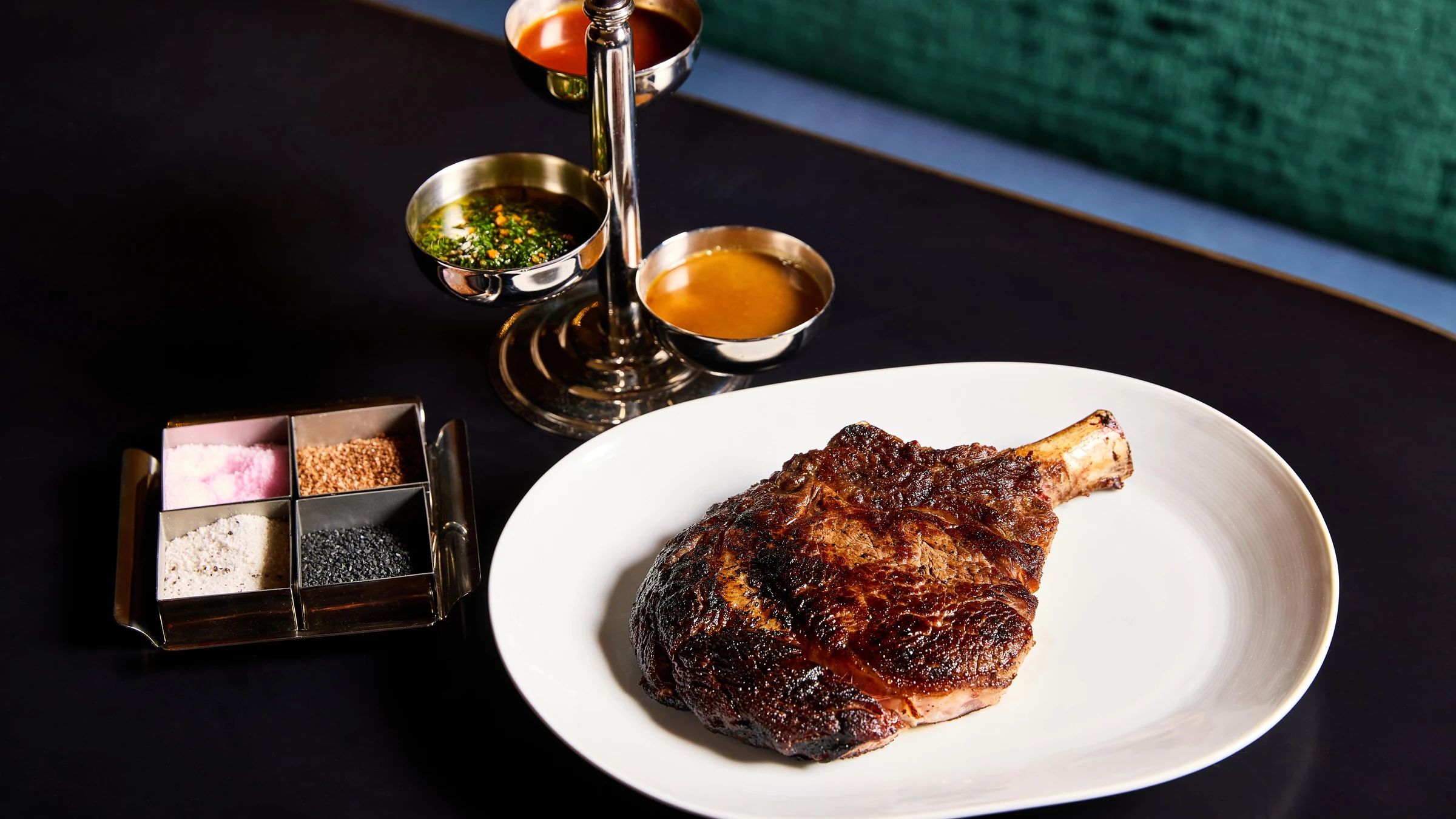 A beautifully plated bone-in ribeye (cowboy steak) with charred crust on a white plate, accompanied by a tiered stand holding sauces and finishing sal