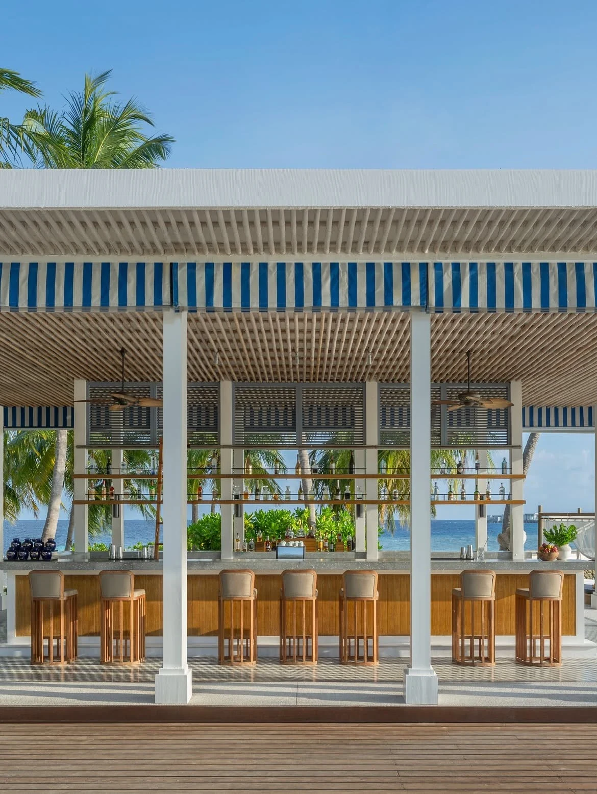 An open-air beachfront bar with wooden bar stools, concrete counter, slatted wooden ceiling, blue-and-white striped awning, palm trees, and ocean view