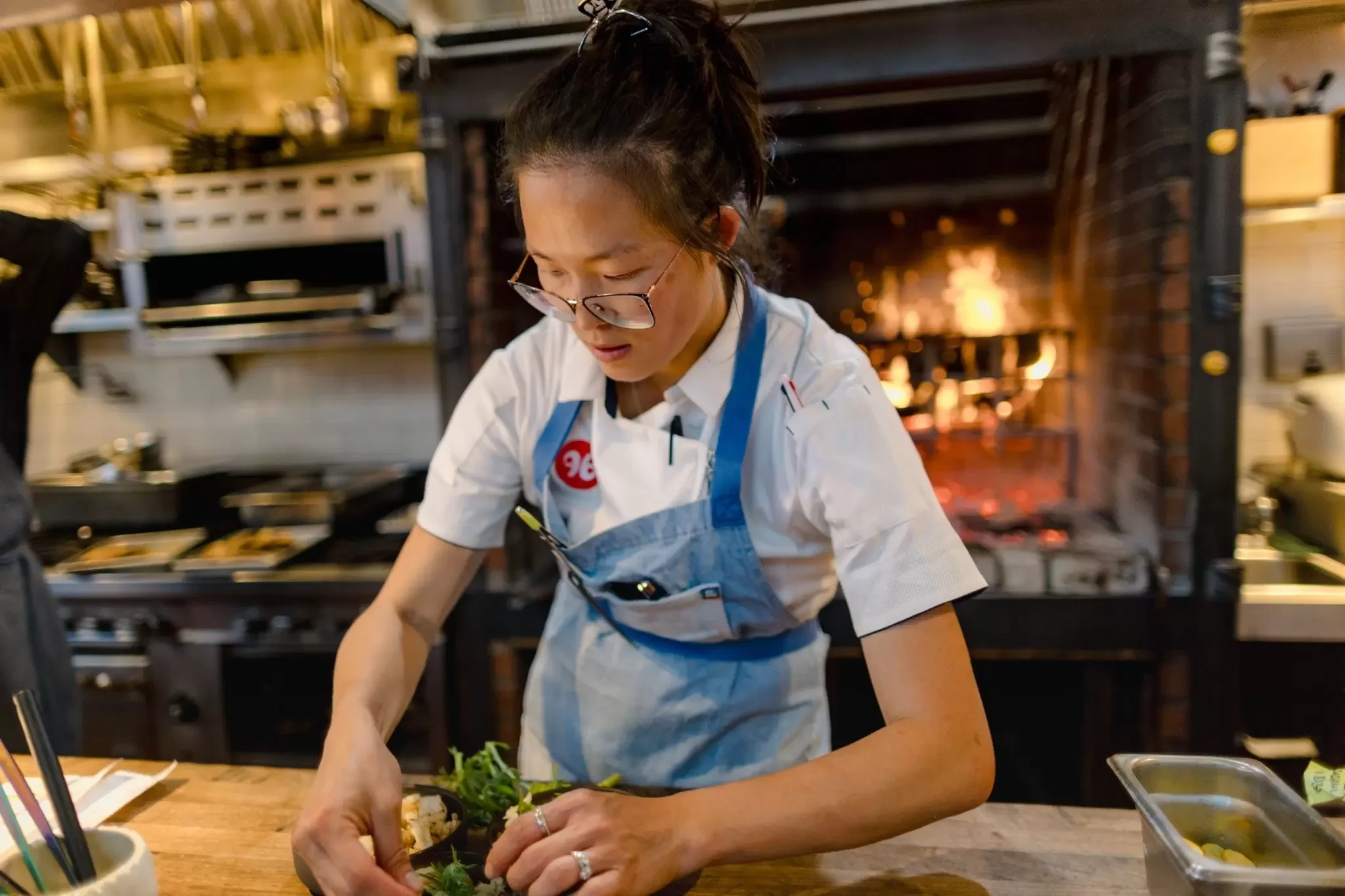 A chef in a blue apron and white shirt with glasses is plating food in a professional kitchen with a wood-fired oven visible in the background; the so