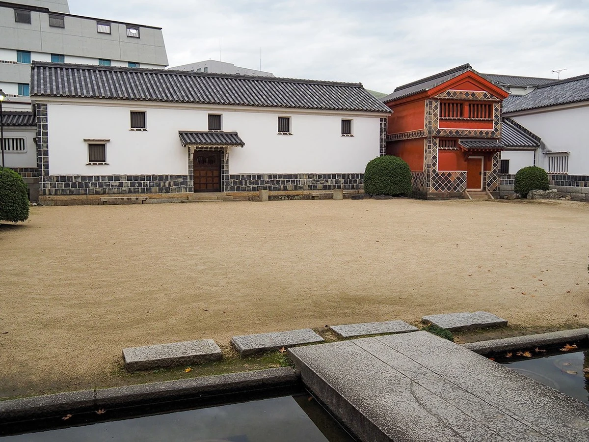 Exterior courtyard of traditional Japanese buildings with white walls, dark tiled roofs, and a distinctive red-painted structure, surrounded by