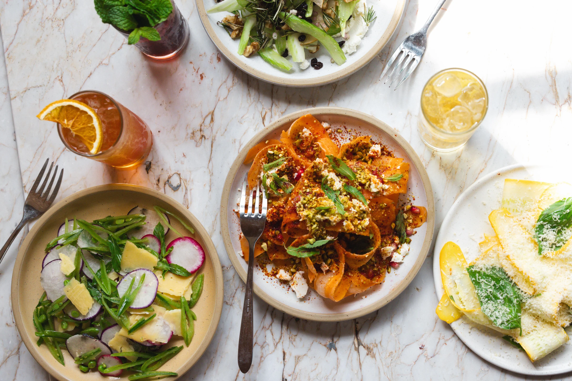Overhead shot of multiple Italian dishes including pasta with vegetables, a fresh salad with radishes, and cocktails on a marble table.