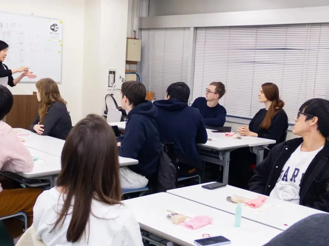 An instructor gestures at a whiteboard with Japanese text while a diverse group of students at individual desks listen attentively in a classroom.