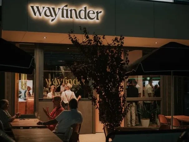 The illuminated "wayfinder" sign above the entrance of the Wayfinder venue at night, with patrons seated at outdoor tables and visible inside.