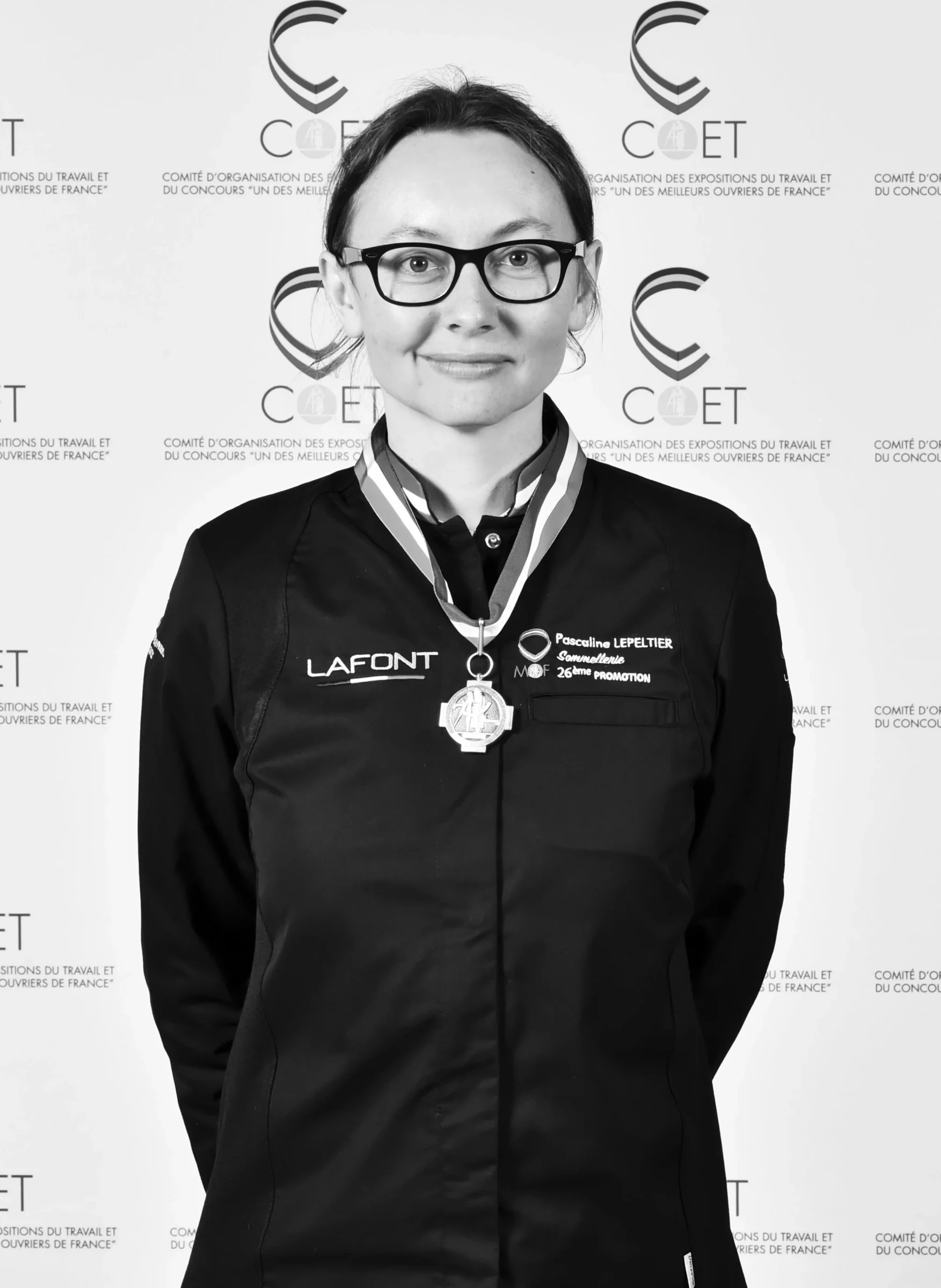 A woman wearing glasses and a black chef's jacket with 'LAFONT' branding and 'Pascaline LEPELTIER' embroidered on it, wearing a medal on a ribbon arou