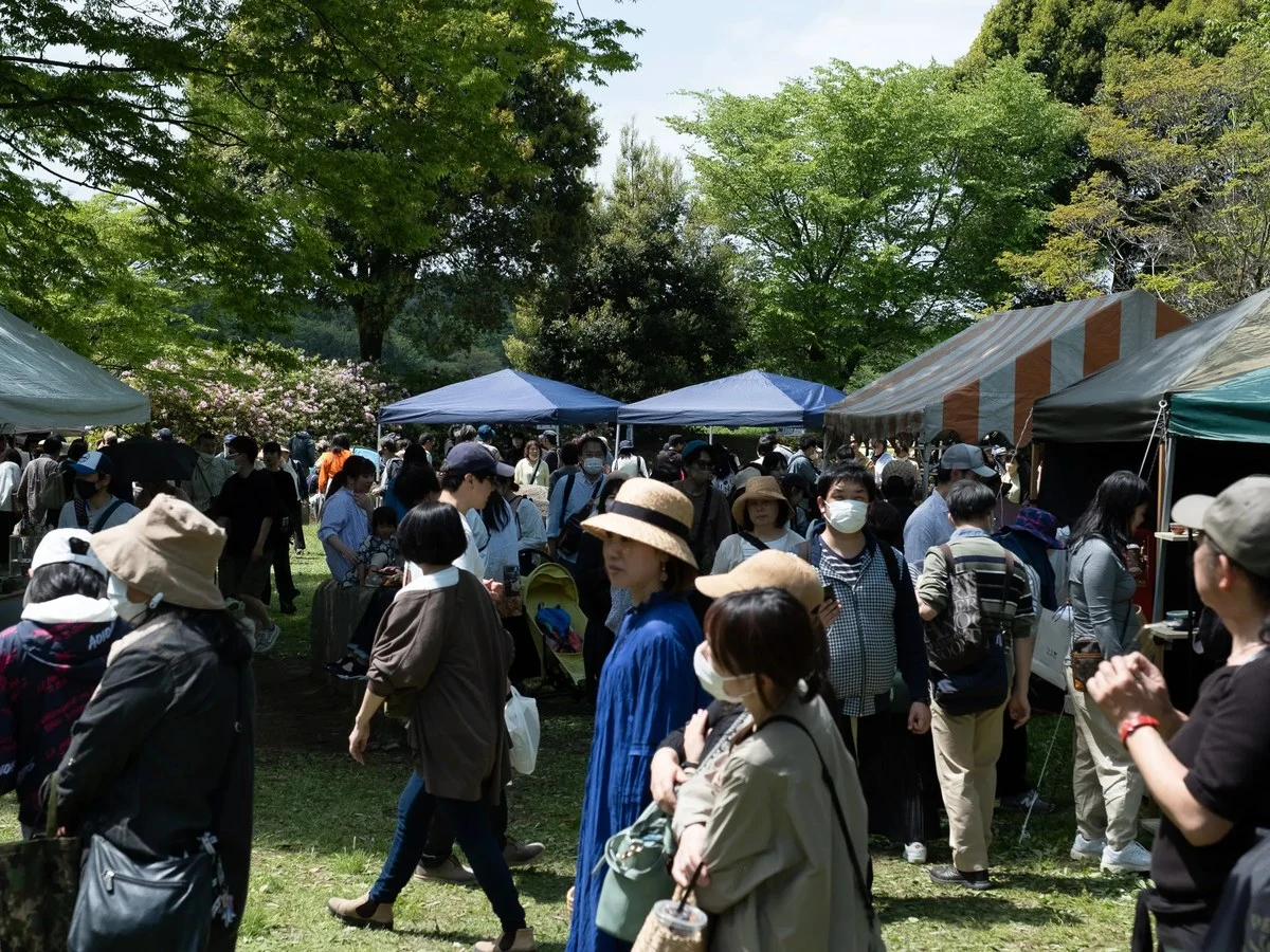 A large crowd of people walks through an outdoor market with white and blue canopy tents, surrounded by green trees.
