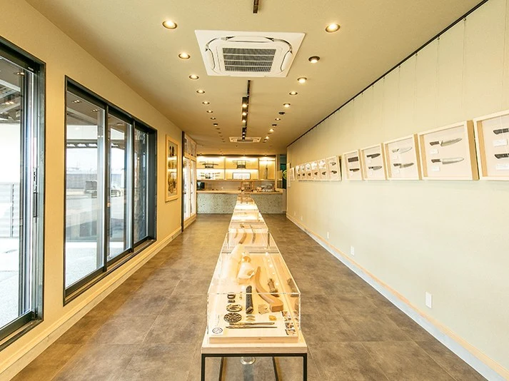 Interior of a long, narrow gallery with glass display cases showing blades and cutlery, framed knife illustrations on the right wall, and a service