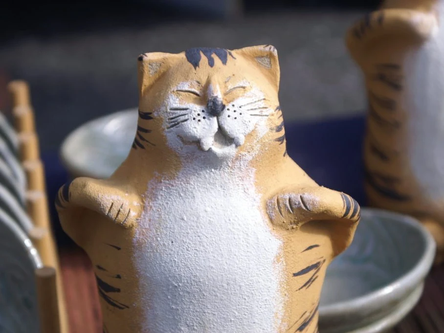 A ceramic cat figurine, orange and white with black stripes, smiles with closed eyes, surrounded by pottery bowls and dishes.