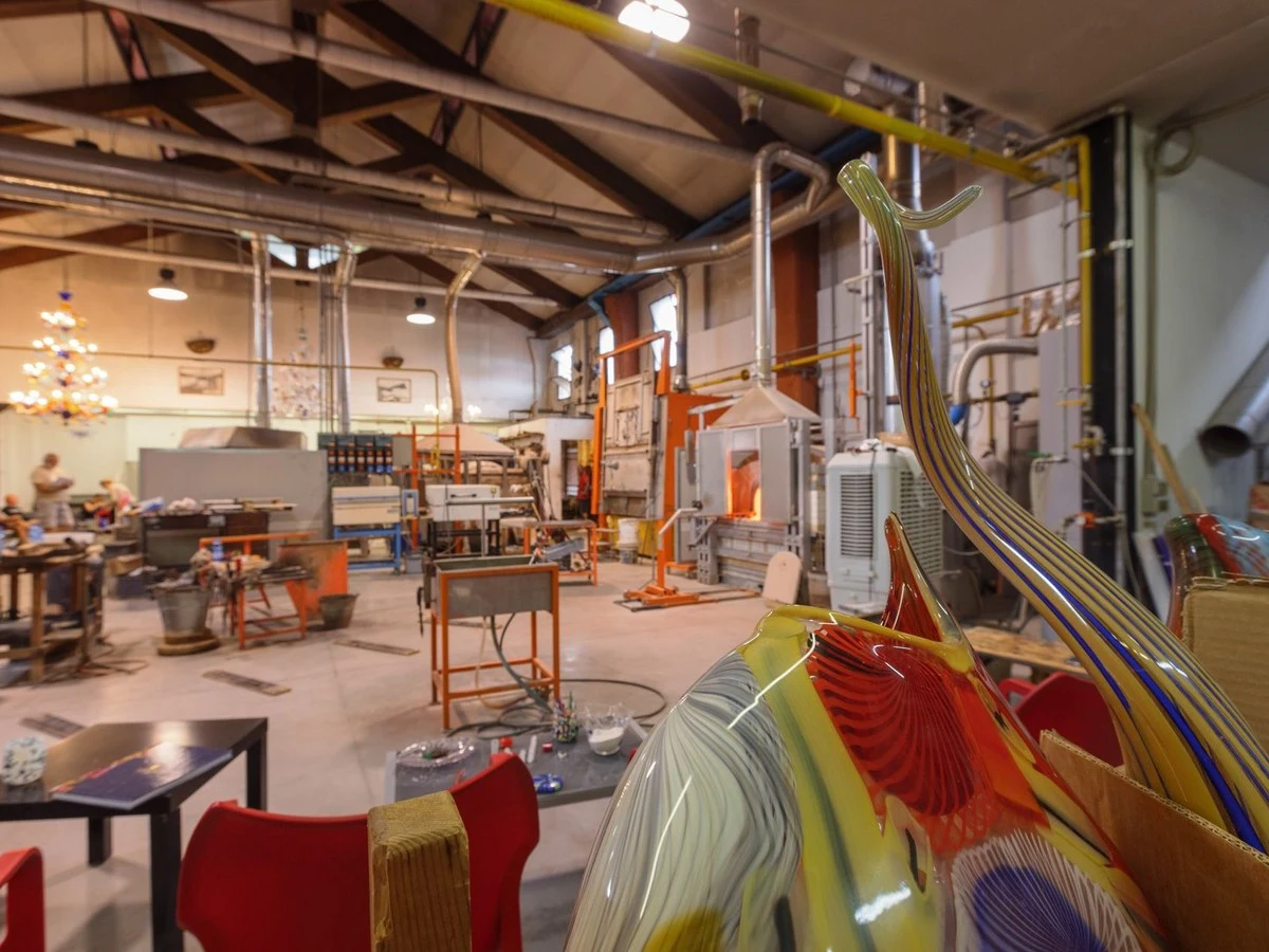 A Murano glass workshop with a glowing furnace, tools, and a colorful glass sculpture in the foreground.