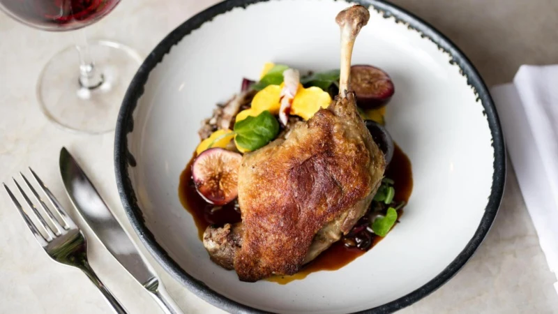 A plated duck confit dish with crispy skin, served with vegetables and sauce on a white plate with black rim, published by Resy in an article explicit
