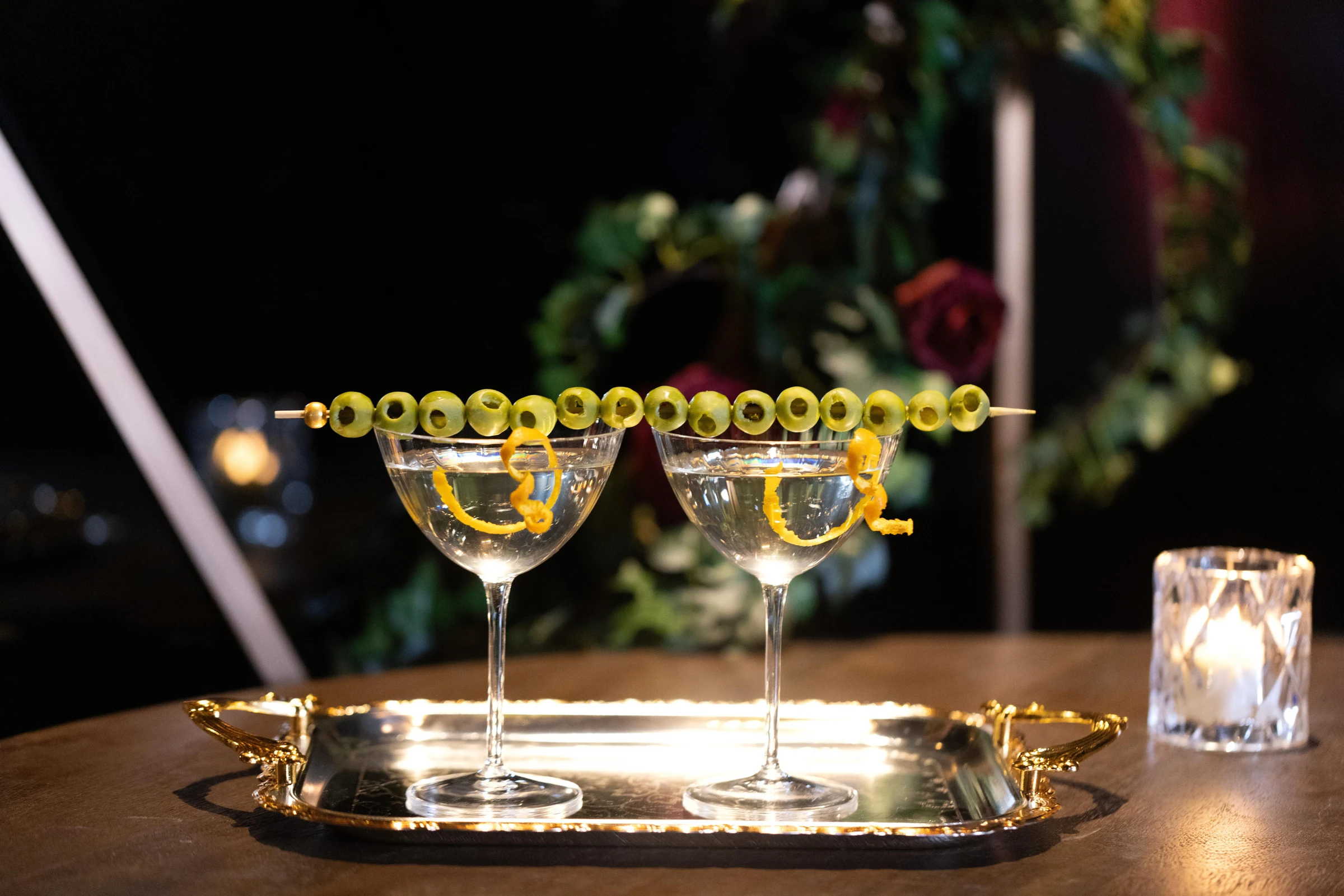 Two elegant coupe cocktails garnished with olives and lemon twists on a gold serving tray in an upscale bar or restaurant setting.