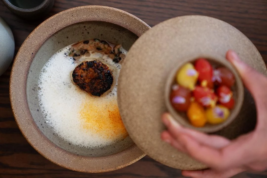 A fine-dining tasting menu course featuring a charred protein element in a foamy broth with colorful heirloom tomatoes in a separate bowl, presented o
