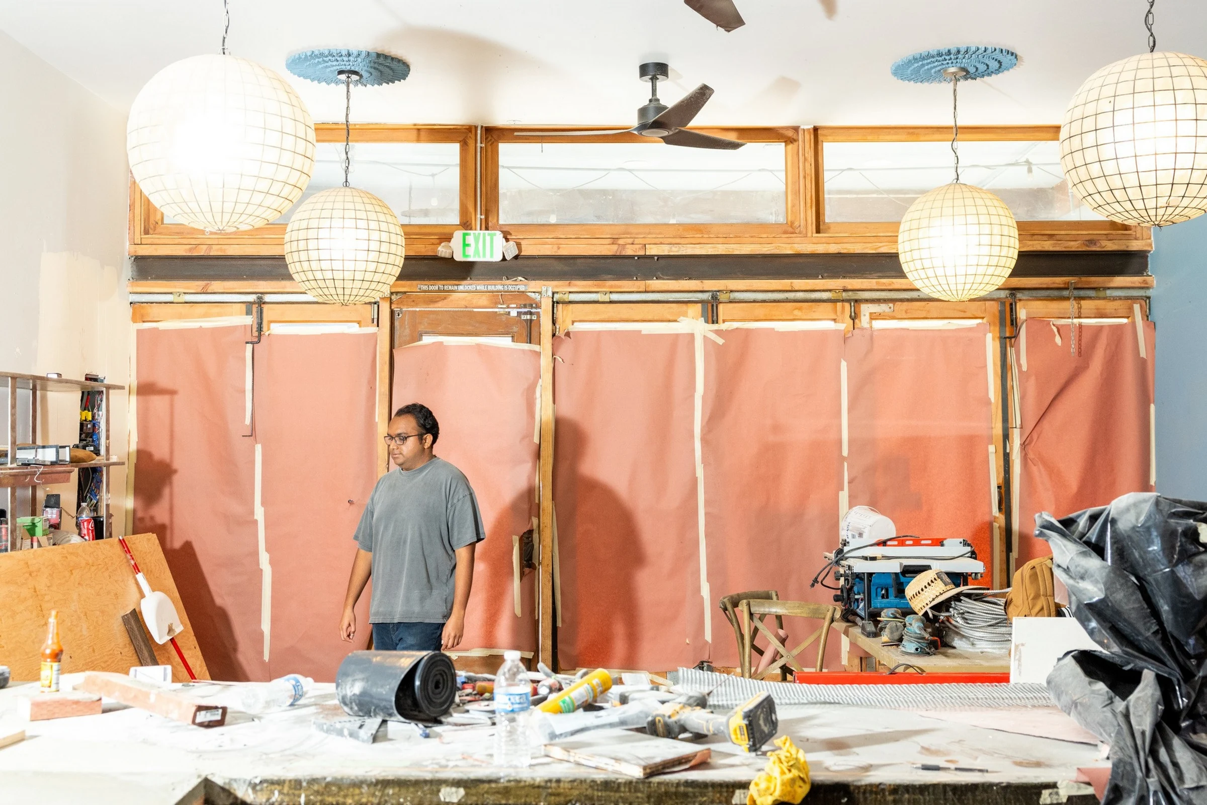 A person stands in a construction/renovation space with exposed framing, pink insulation sheets, hanging paper lanterns, and scattered tools and mater