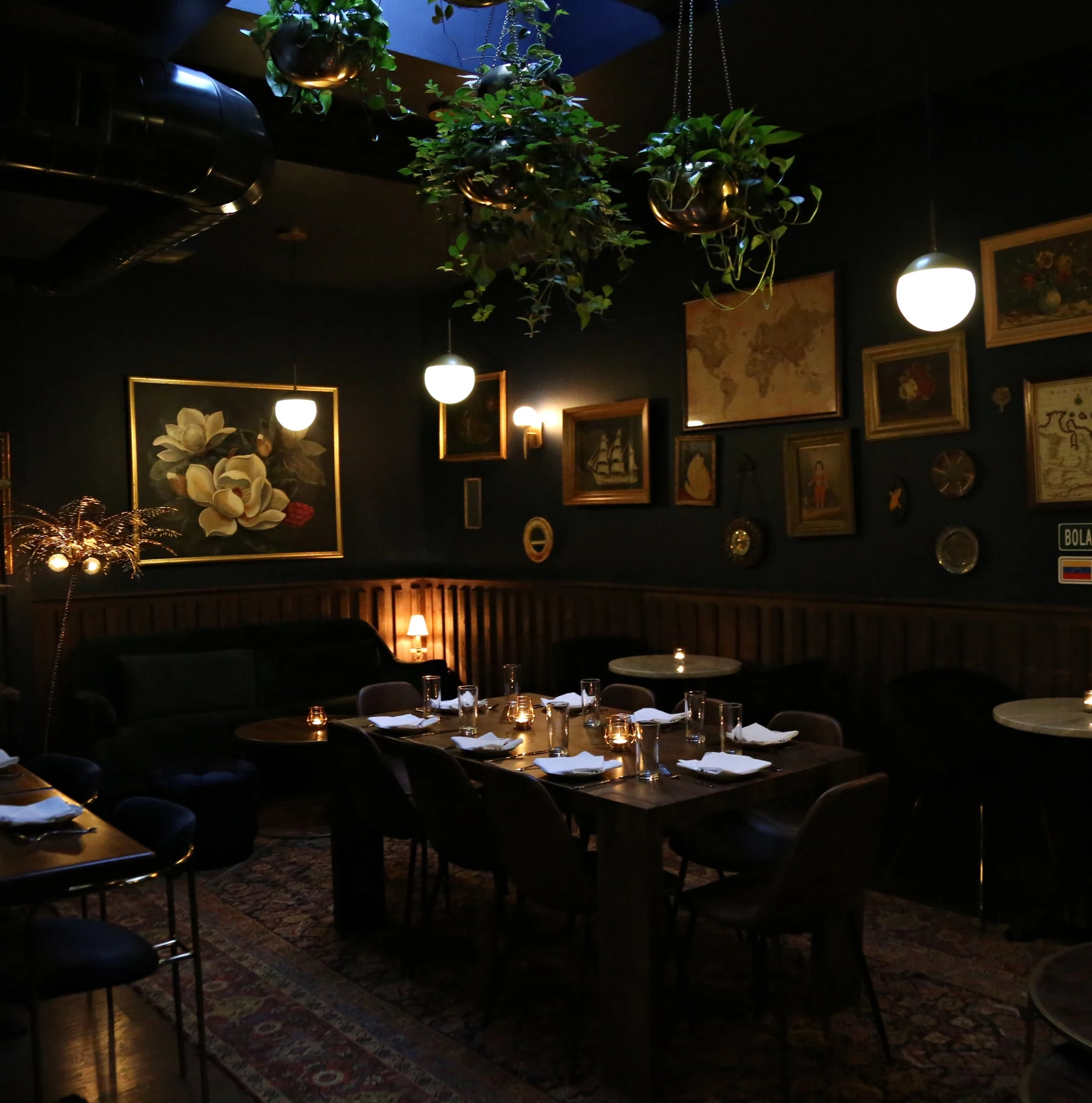 An upscale restaurant dining room interior with dark walls, eclectic framed artwork, globe pendant lights, hanging plants, and set tables with white n