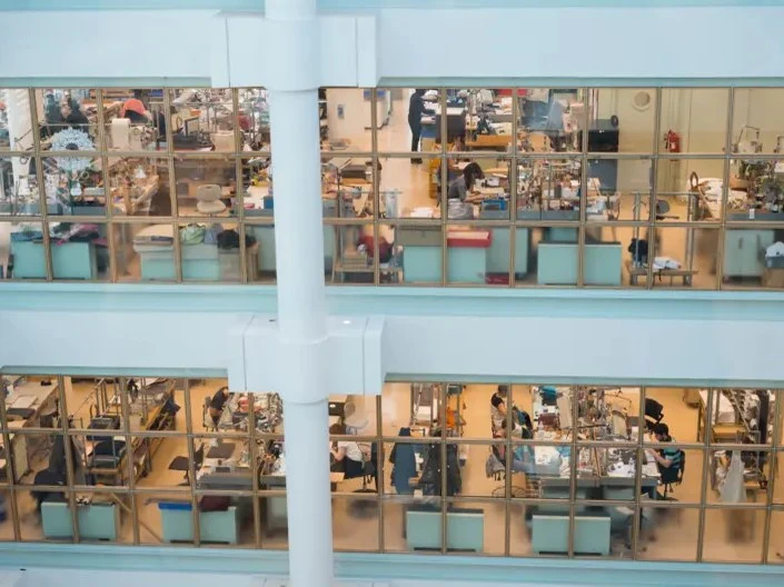 A multi-story workshop with numerous artisans working at individual benches, visible through large glass windows, consistent with an Hermès atelier.