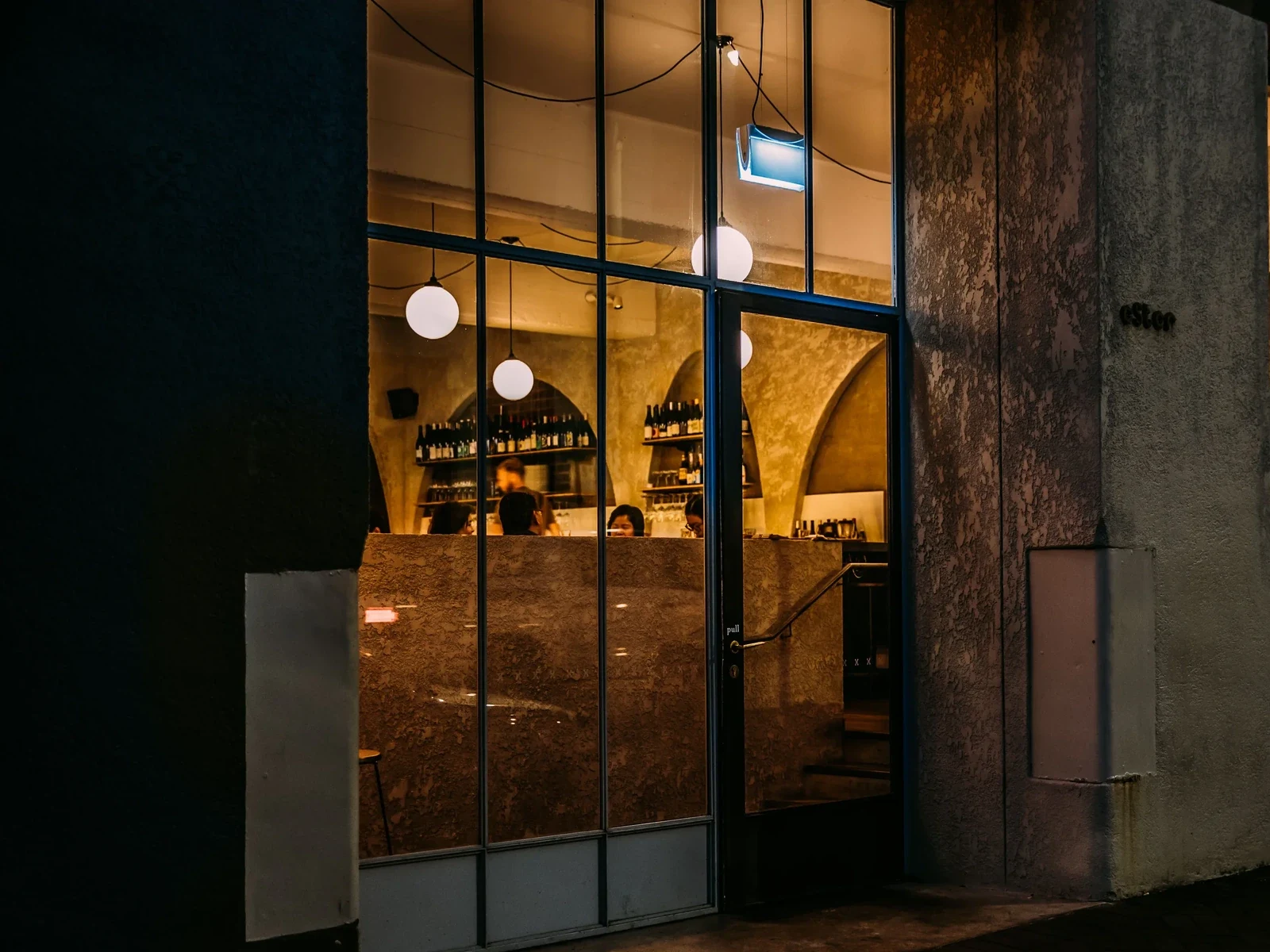 A nighttime exterior view of a wine bar with black-framed glass windows, warm interior lighting, hanging globe lights, and wine bottles visible on shelves behind the counter.