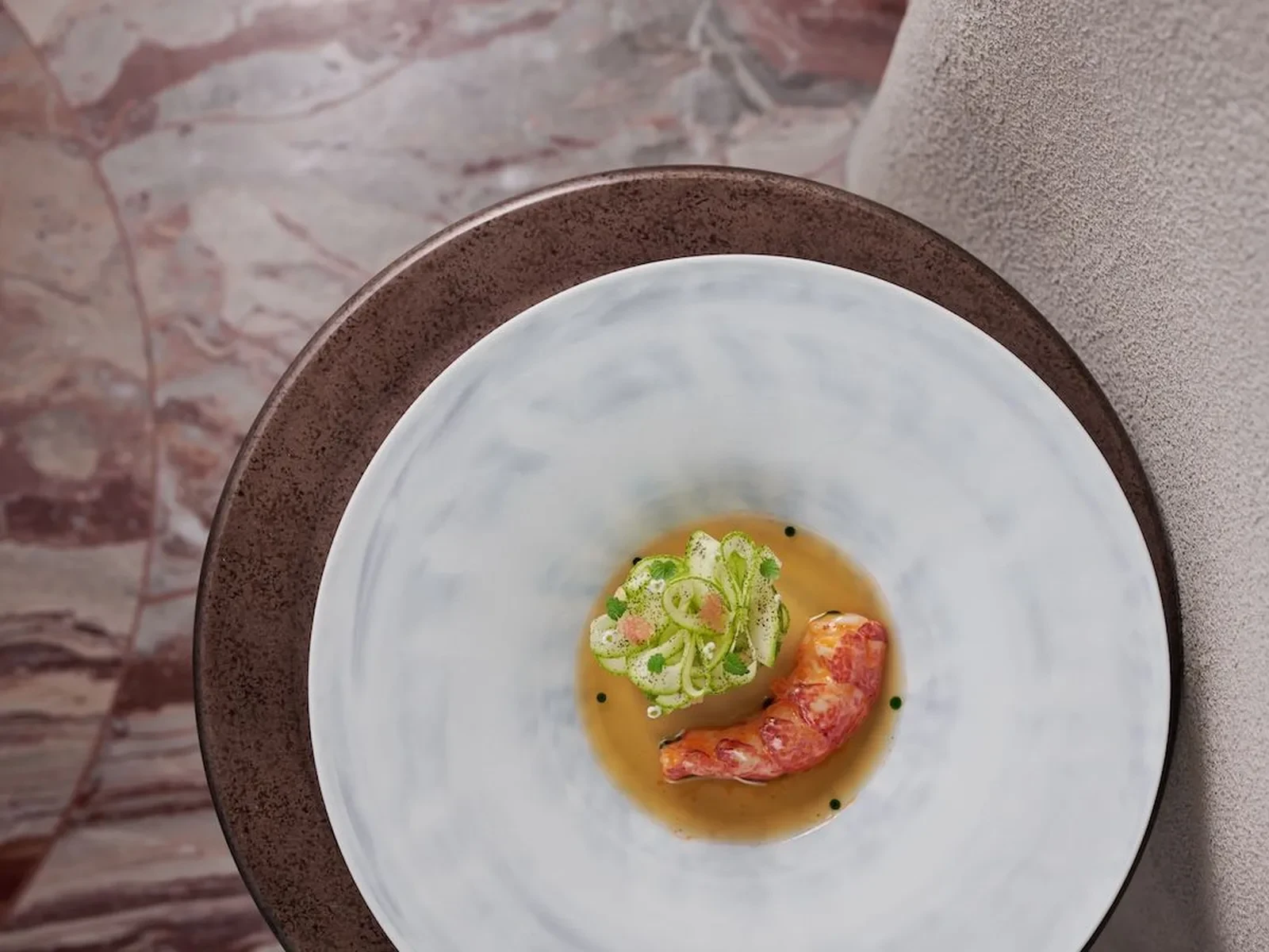 Dish featuring lobster, green garnishes, and broth on a marble table.