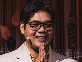 A man in a white suit jacket with glasses smiles and makes a prayer gesture on a stage with bottles of Johnnie Walker Black Label.