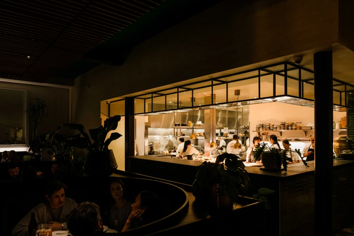 An upscale restaurant interior at night showing an open kitchen with chefs working, diners seated in the foreground, and modern architectural elements