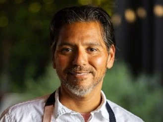 A man with dark hair and a beard, wearing a white shirt and an apron, smiles at the camera.