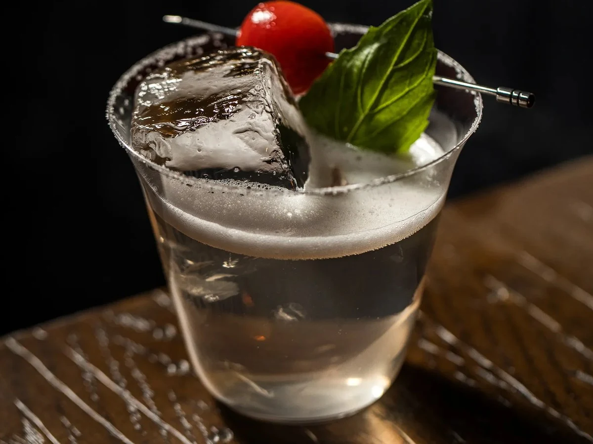 A craft cocktail in a rocks glass with a large clear ice cube, garnished with a cherry tomato and fresh basil leaves on a cocktail pick, served.