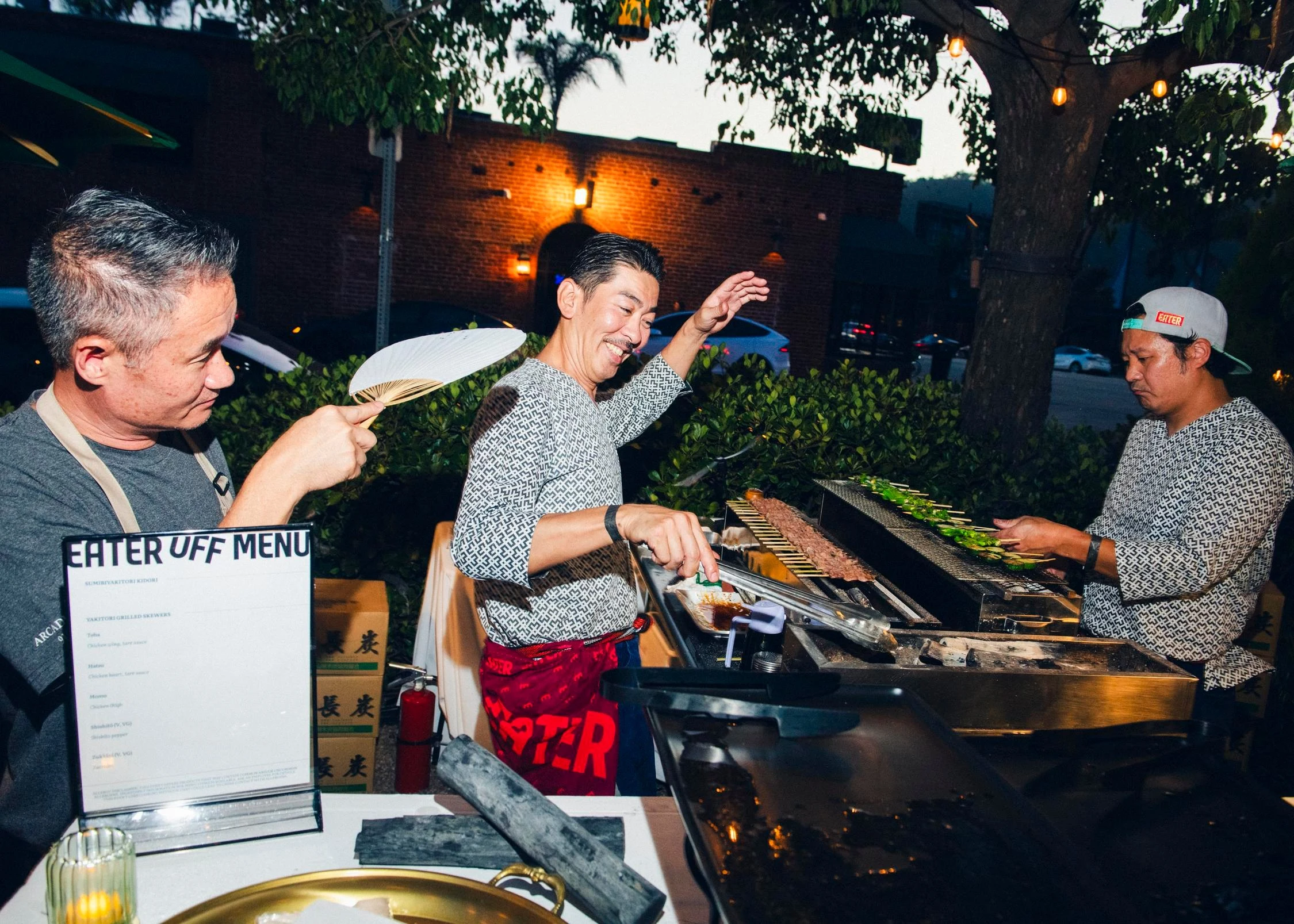 Three men working at an outdoor grilling station at dusk, with one man fanning the grill and two others tending to skewers and meat