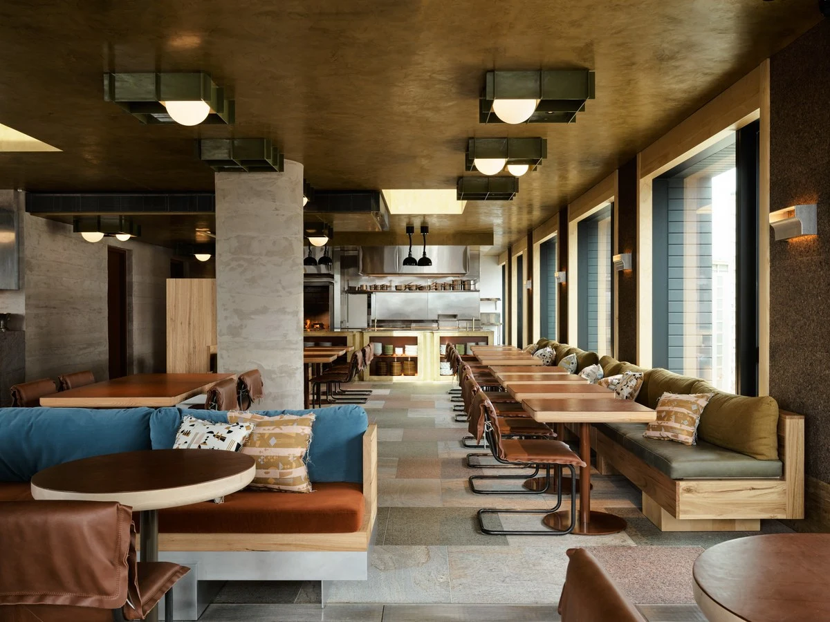 Modern restaurant interior with a gold-colored ceiling, concrete columns, booth seating, and an open kitchen.