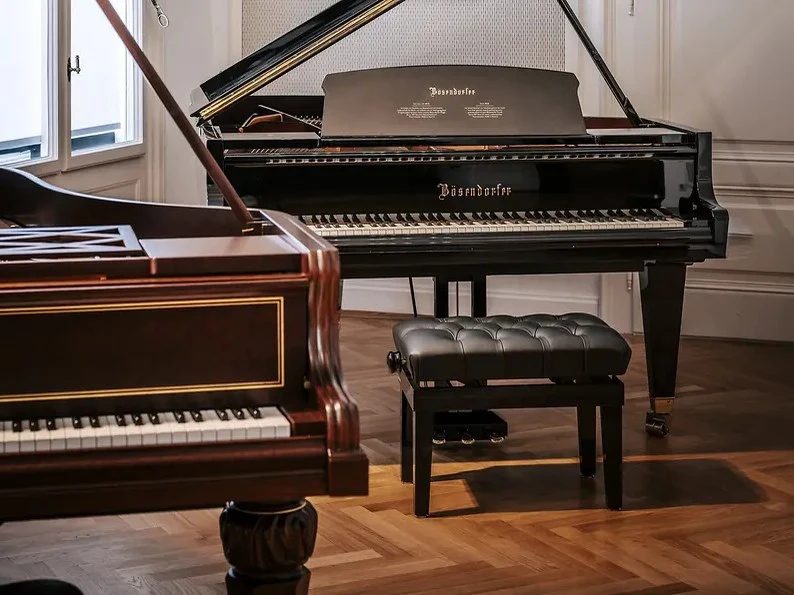 Two grand pianos, one dark wood and one black Bösendorfer, with a tufted leather bench on a herringbone floor in a showroom.