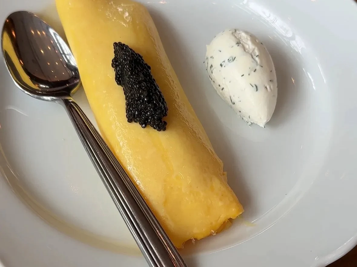 A rolled French omelet topped with black caviar, served alongside a quenelle of herbed crème fraîche on a white scalloped plate with a spoon.