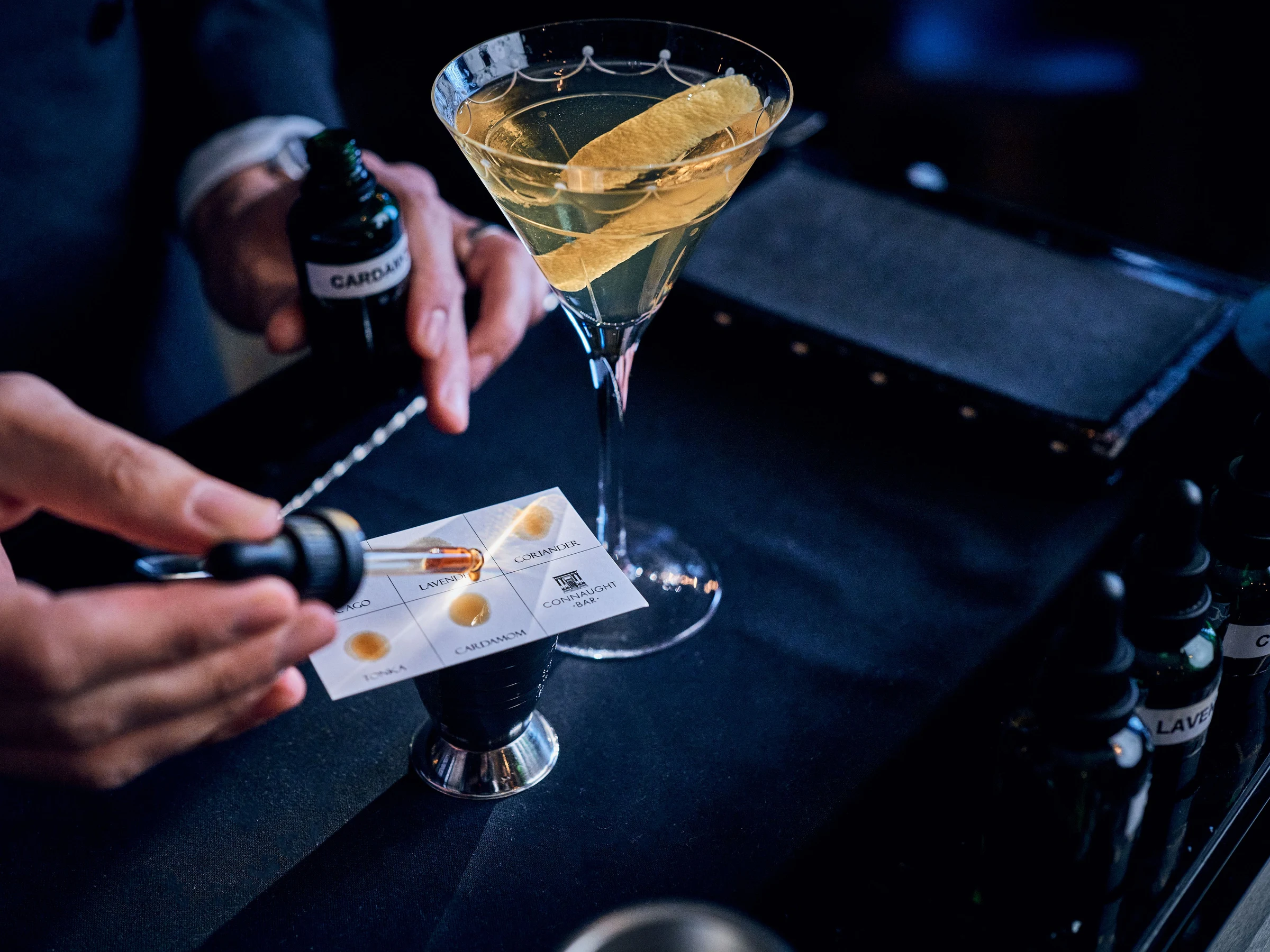 A bartender's hands hold a bottle and tasting card next to a martini glass filled with golden liquid and a lemon twist on a dark bar counter.