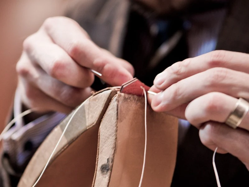 Close-up of hands performing precise hand-stitching on a tan leather piece using needle and thread.