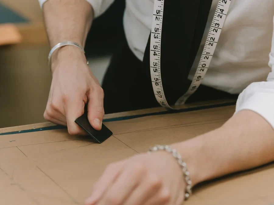 A tailor from Huntsman Savile Row uses a chalk block to mark a pattern on brown paper, with a measuring tape draped around their neck.