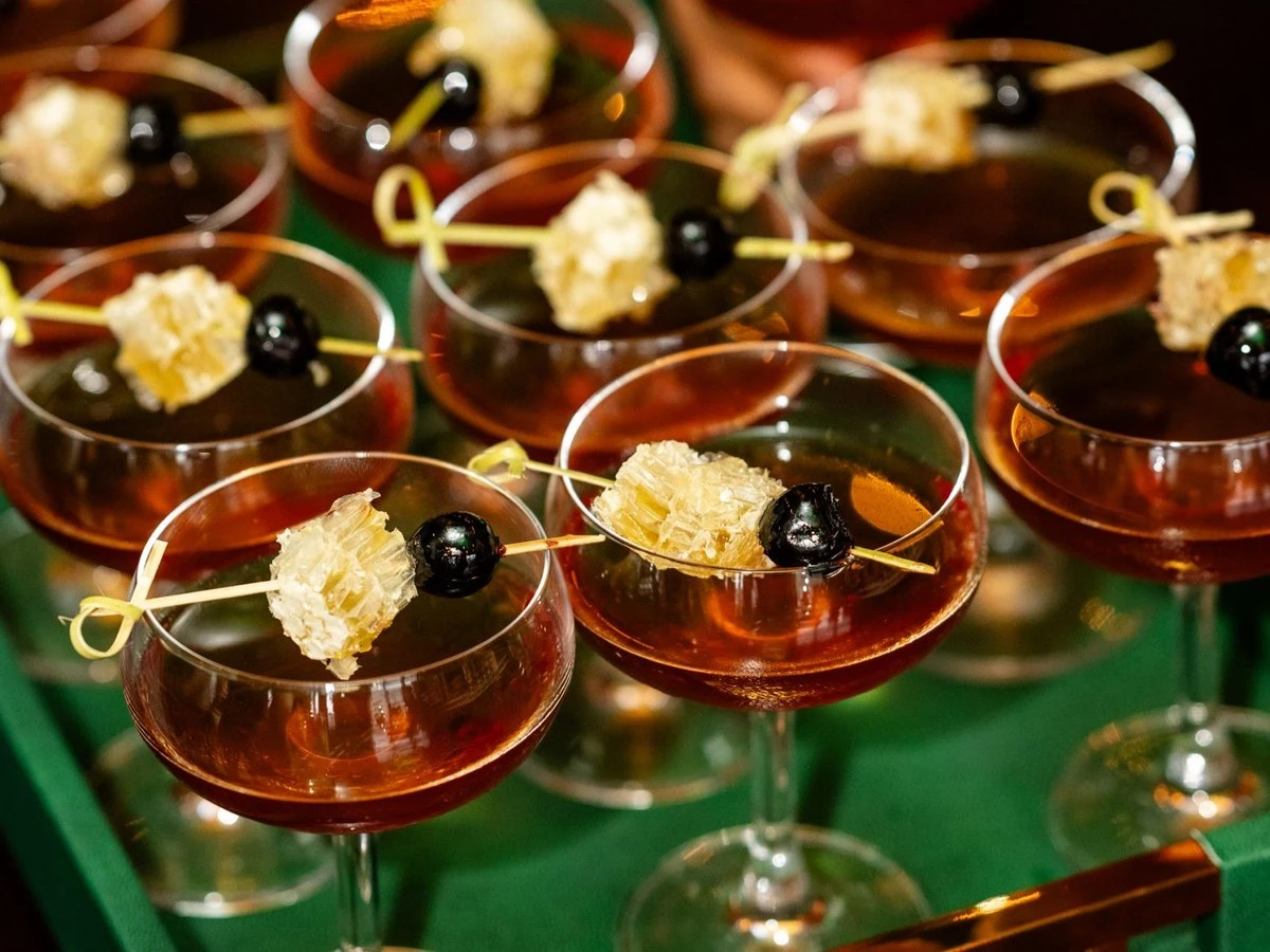 Multiple coupe glasses filled with amber cocktails, each garnished with a piece of honeycomb and a black cherry on a decorative pick, arranged.
