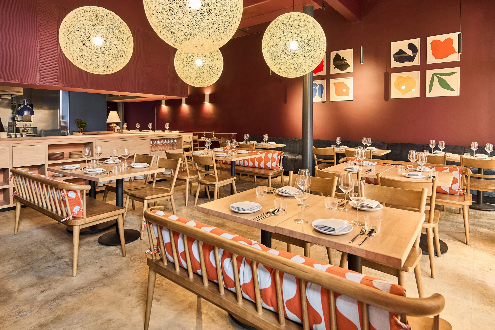 A modern restaurant dining room with natural wood tables, terracotta-burgundy walls, sculptural spherical pendant lighting, and colorful abstract art;