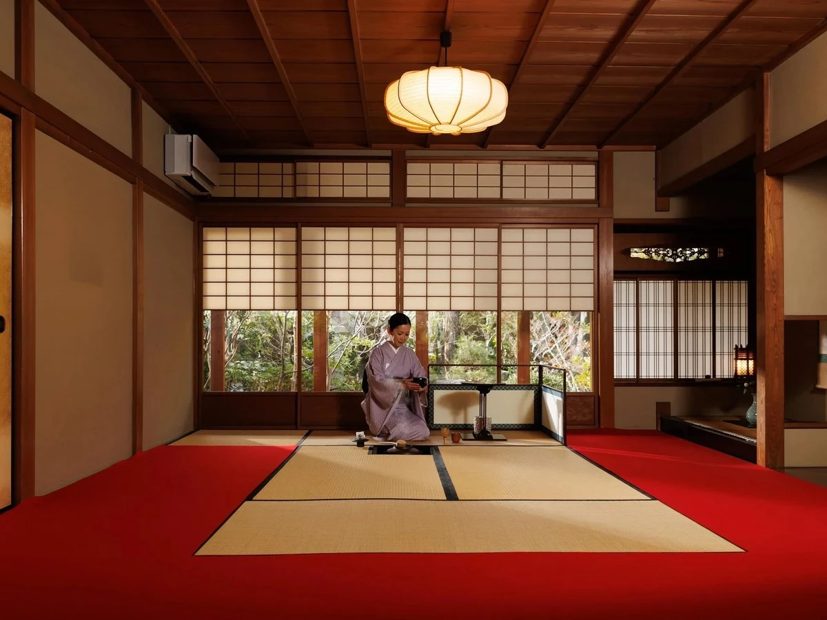A woman in a lavender kimono kneels on tatami mats in a traditional Japanese room with shoji screens, a wooden ceiling, and a paper lantern.
