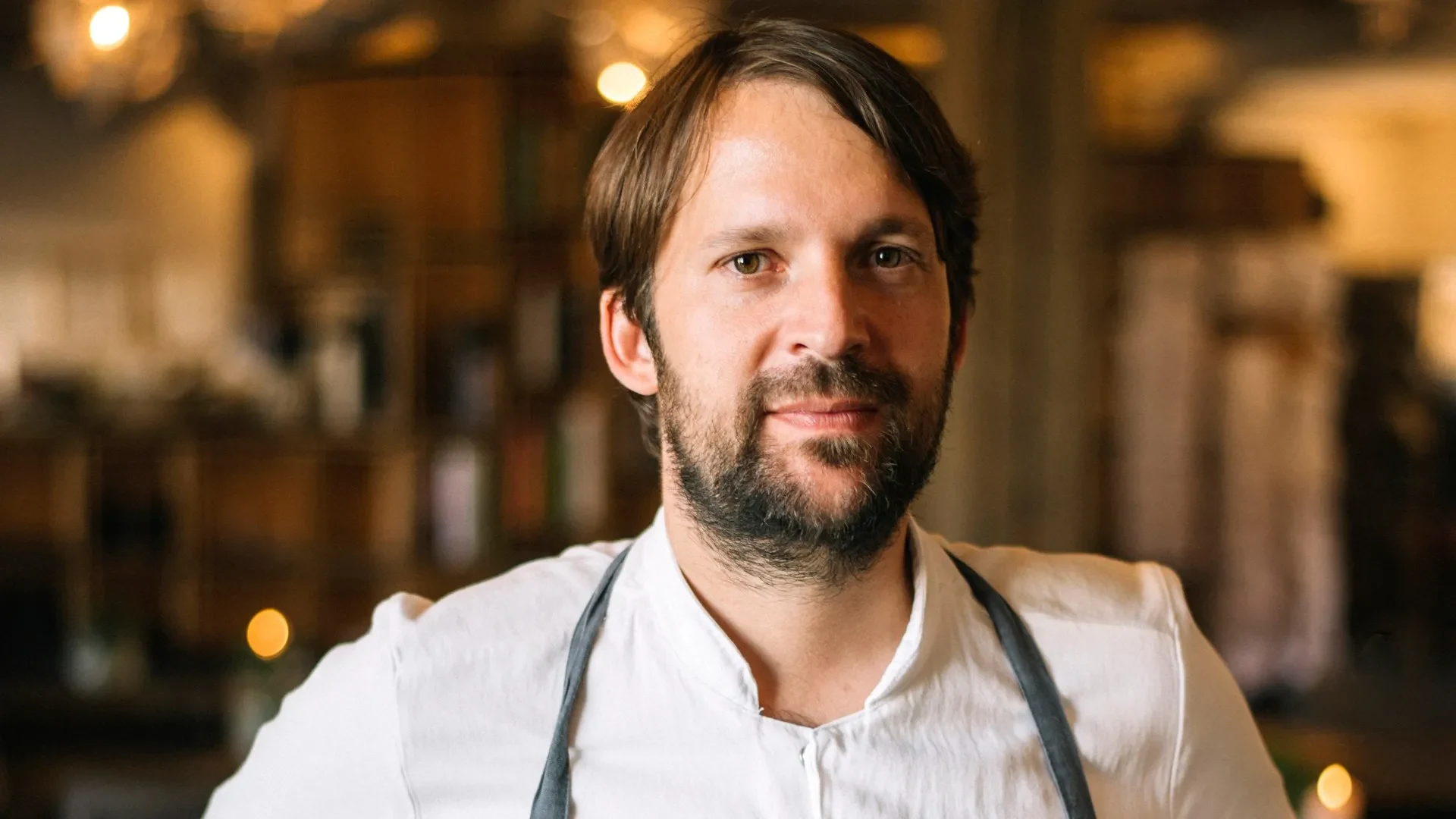 A man with a beard and dark hair, wearing a white shirt and a grey apron, looks directly at the camera.