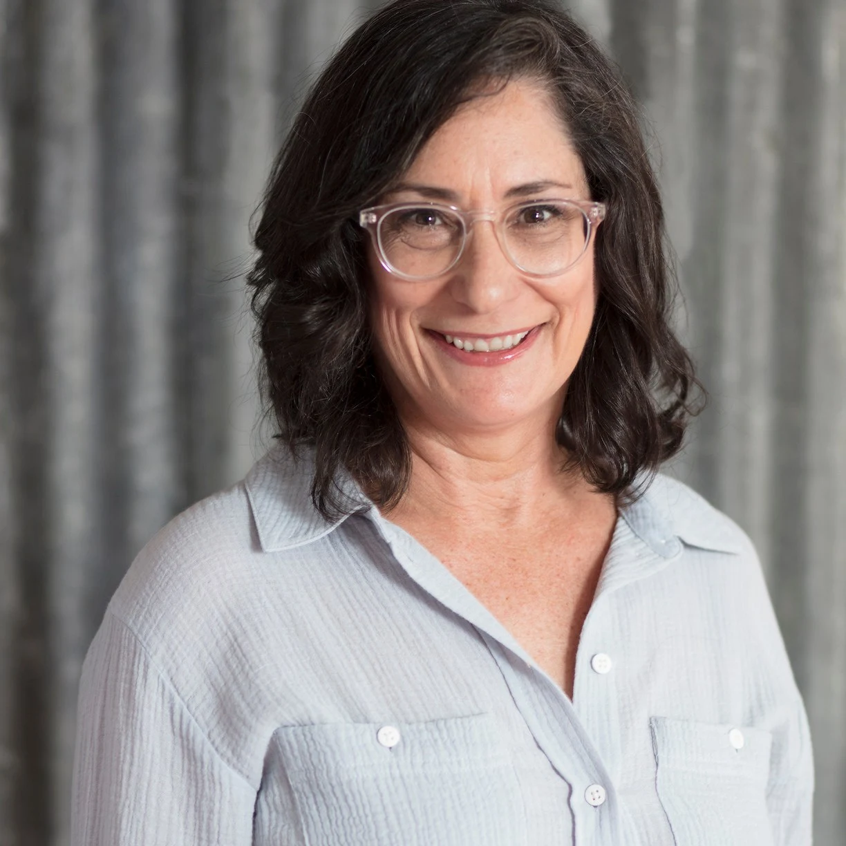Professional portrait of a woman with dark wavy hair, clear-framed glasses, and a light blue button-up shirt against a neutral background