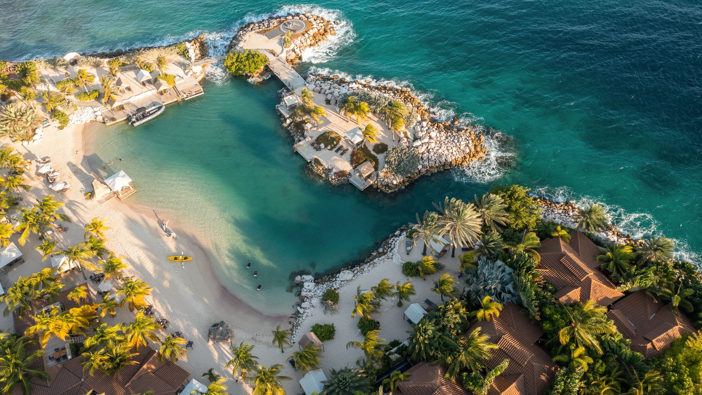 Aerial view of a luxury beachfront resort with villas, palm trees, white sand beach, and turquoise water with rocky outcroppings and walkways; source