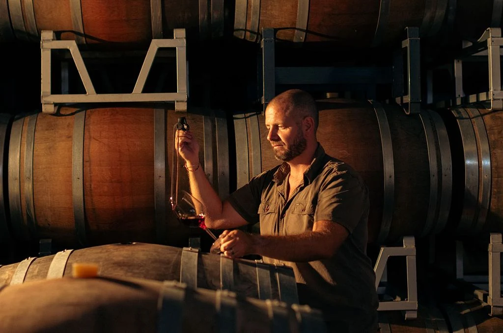 A man in a short-sleeved work shirt examines a wine glass in a barrel cellar